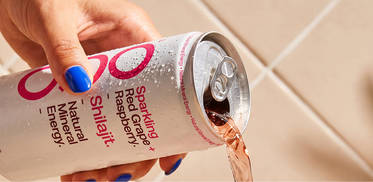 A hand with blue nail polish holds a sparkling red beverage can as pinkish-red liquid pours out into a tiled background.